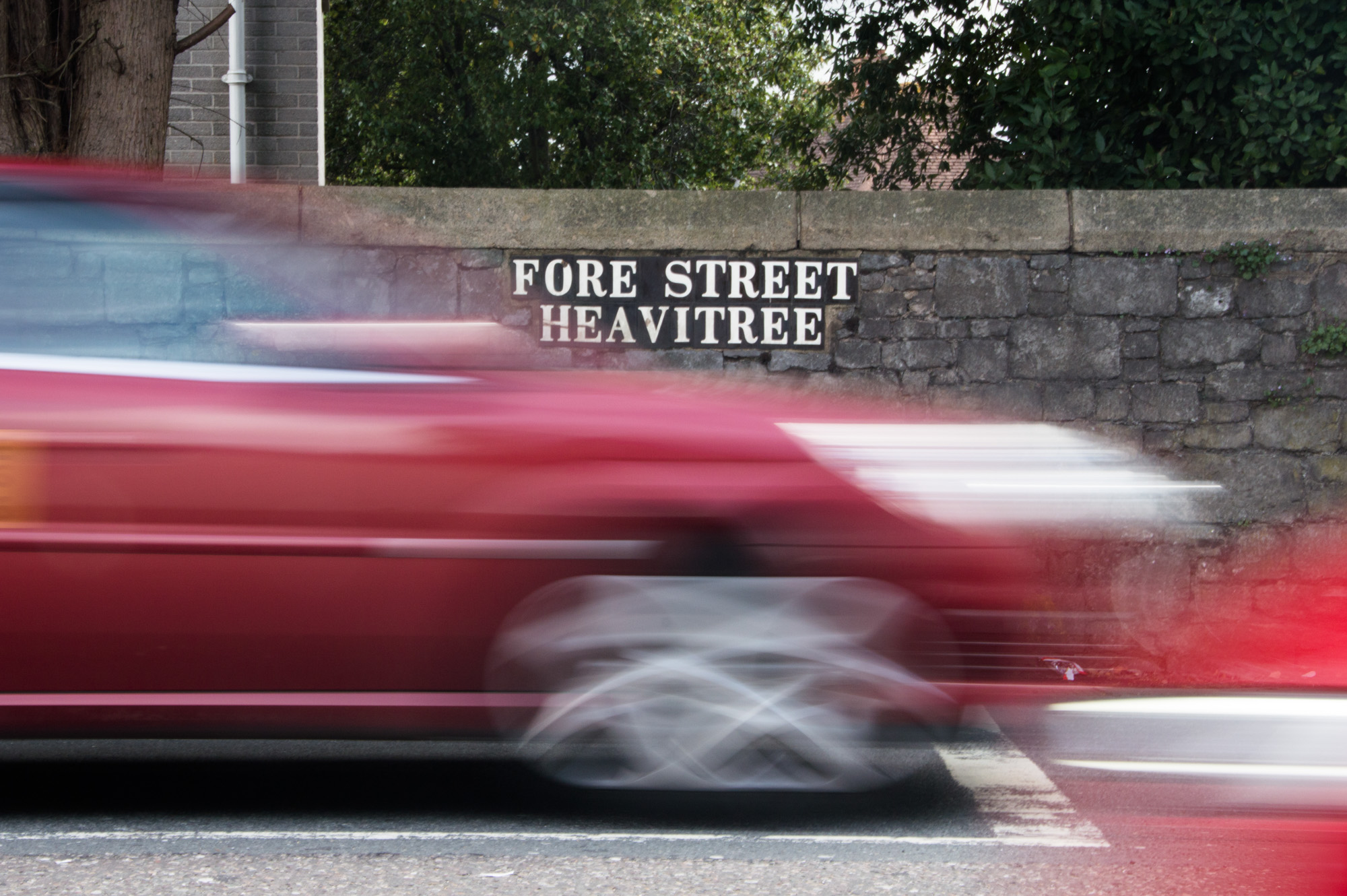 An image of the street name Fore Street Heavitree written in bold white with an image of a red car in motion going past in the foreground